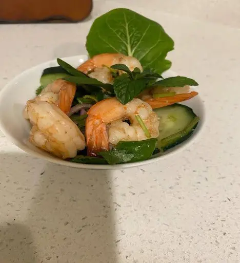 A bowl of shrimp salad with cucumber slices, green peppers, purple onions, and a leafy green garnish on a white counter.