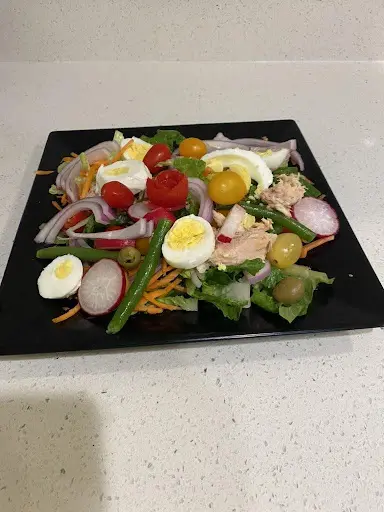 A colorful salad on a black square plate, featuring mixed greens, cherry tomatoes, red onions, sliced radishes, shredded carrots, boiled eggs, and chunks of chicken.