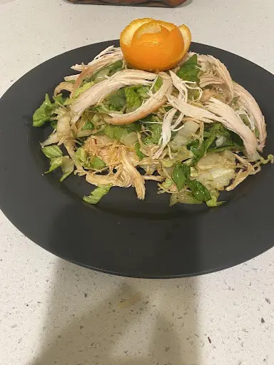 A plate of shredded chicken salad with lettuce, herbs, and a decorative orange peel on top, served on a black plate against a white countertop.