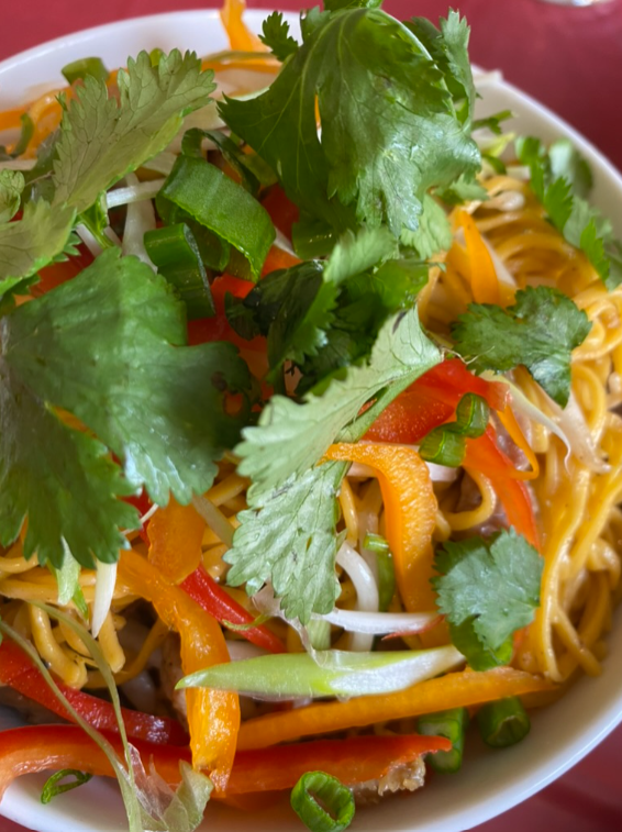 A bowl of asian noodles topped with fresh cilantro, sliced bell peppers, and bean sprouts, served on a red table.